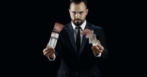 Professional Young Magician in a Dark Suit Throws Two Jets of Cards into the Air in Slow Motion. Background is Black.  - Video