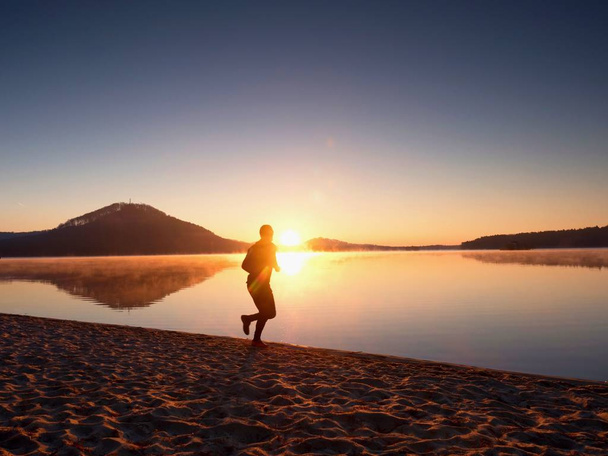 Man running on beach against backdrop of a beautiful sunset. Sand of mountain lake - 写真・画像