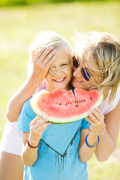 A blond boy holds a watermelon slice in his hands. The boy is eating a watermelon. Mom and son laughing - Photo, Image