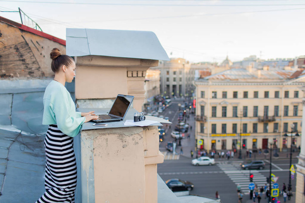 schöne und stilvolle Mädchen arbeitet für einen Laptop auf dem Dach des Hauses in der Altstadt. Auf dem Tisch liegen auch Dokumente und eine alte Kamera - Foto, Bild