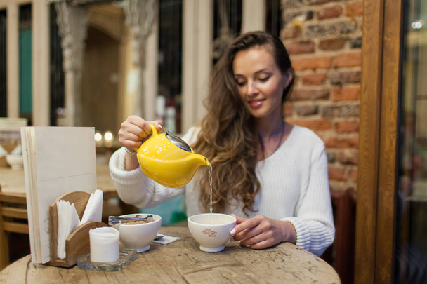Smiling young girl in the restaurant pours green tea from the yellow teapot in a Cup. Focus on a mug of tea - Photo, Image