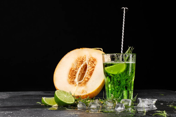 A green drink with lime and tarragon. A cocktail and a melon on a black background. Healthy, sweet and tasty drinks. Copy space. - Фото, изображение