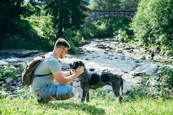 joven hombre fuerte se queda cerca de las montañas río con perro y en busca de la vista
 - Foto, imagen