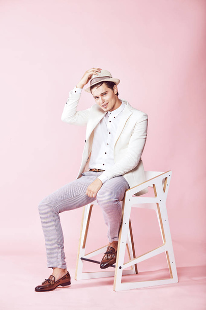Jonge knappe gelukkig lachend moderne man dragen witte jas, wit t-shirt, grijze broek en witte hoed zitten op de stoel in studio met licht roze achtergrond en die van zijn hand op de hoed - Foto, afbeelding
