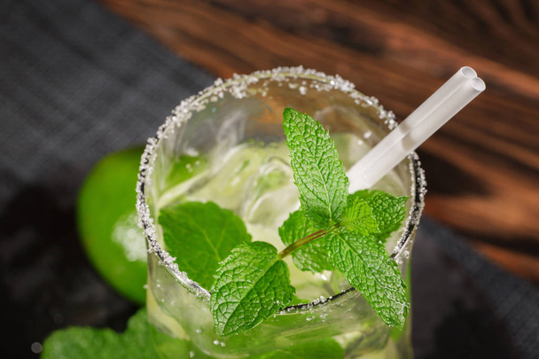 A glass filled with alcohol beverage from juicy lime, rum, fresh mint and crushed ice on a dark wooden background. Summer mojito with straw, top view. - Fotó, kép