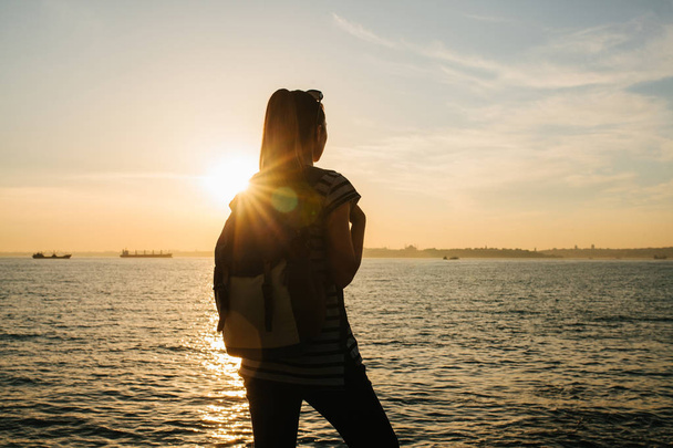 Молода туристична дівчина з рюкзаком стоїть біля моря на заході сонця і дивиться на відстань. Відпочинок, розслаблення, подорожі, відпустка
. - Фото, зображення