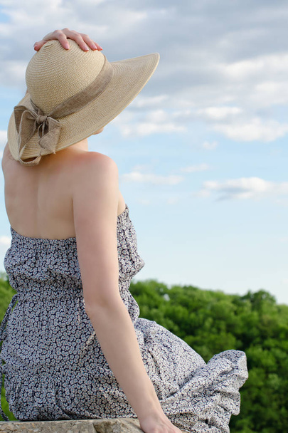 Beautiful girl in hat and dress sits on a background of cloudy blue sky and forest - Foto, afbeelding