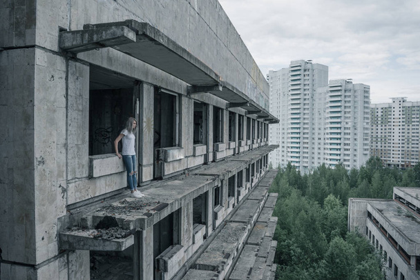 A young girl in a white T-shirt and blue jeans is standing on the edge of a ruined balcony in a ruined building early in the morning. - Photo, image