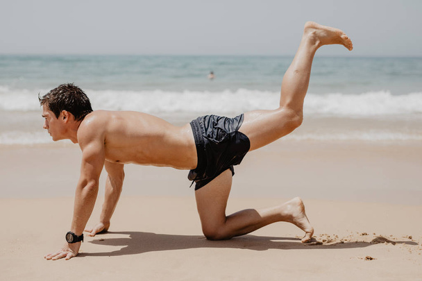 Αθλητής εκτέλεση ασκήσεων στην παραλία κάτω από τον ήλιο. Νεαρός εκπαίδευση στην παραλία - Φωτογραφία, εικόνα