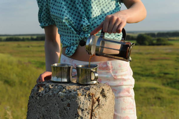 Het meisje giet koffie in twee kopjes uit een Frans-pers in de open lucht. Zomer landschap - Foto, afbeelding