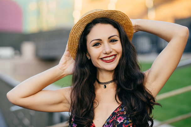 Happy brunette female with pleasant smile wearing hat keeping hand behind head posing outdoors. Pretty woman with happy expression relaxing at big city during her summer holidays. People, emotions - Photo, Image