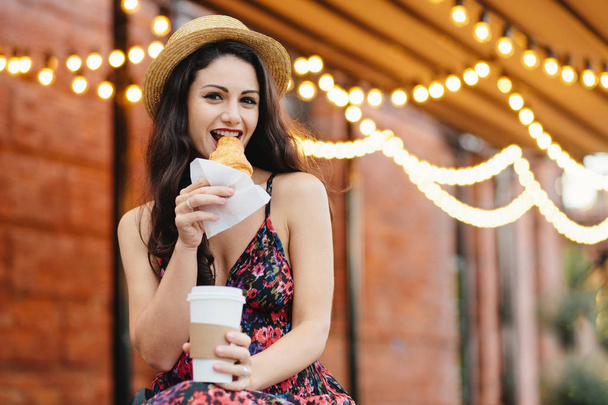 Pretty woman with dark hair, shining eyes eatting with pleasure tasty croissant and drinking takeaway coffee while sitting at cozy balcony. Young woman in summer hat and dress relaxing at cafe - Photo, Image