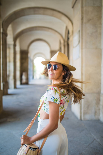 Όμορφη μελαχρινή νεαρή γυναίκα φοράει καπέλο και γυαλιά ηλίου και το περπάτημα σε το δρόμο θερινή ώρα - Φωτογραφία, εικόνα