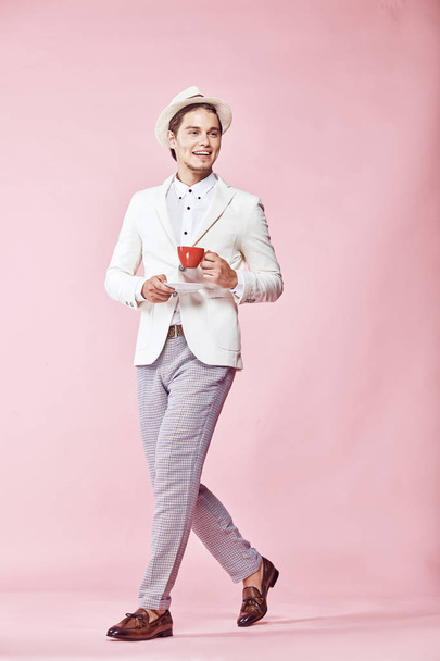 Young beautiful happy smiling modern man wearing white jacket, white shirt, grey pants and white hat stepping through studio with light pink background, holding red coffee cup and looking away - Zdjęcie, obraz