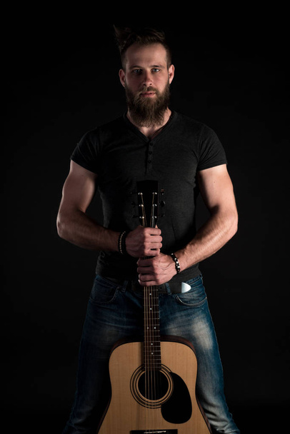 Een charismatische en stijlvolle man met een baard staat met een akoestische gitaar op een zwarte achtergrond van de geïsoleerde full-length. - Foto, afbeelding