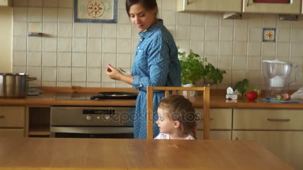 Mom cooks eggs on the stove. A little daughter is waiting for breakfast at the table. The girl is very angry. Childish whims. Family breakfast. - Filmati, video