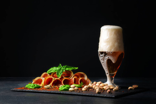 A big glass full of light beer with foam, delicious prosciutto, and spinach on the black background. Pistachios near the alcohol drink. Salty snacks for beer. - Photo, Image