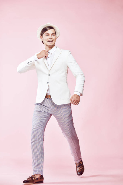 Young beautiful happy smiling modern man wearing white jacket, white shirt, grey pants and white hat stepping in studio with light pink background  - Zdjęcie, obraz