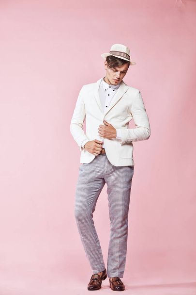 Young beautiful happy serious modern man wearing white jacket, white shirt, grey pants and white hat standing in studio with light pink background and looking down - Fotografie, Obrázek