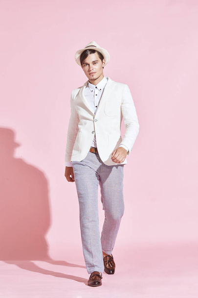 Young handsome serious modern male model wearing white jacket, white shirt, grey pants and white hat stepping in studio with light pink background - Фото, зображення