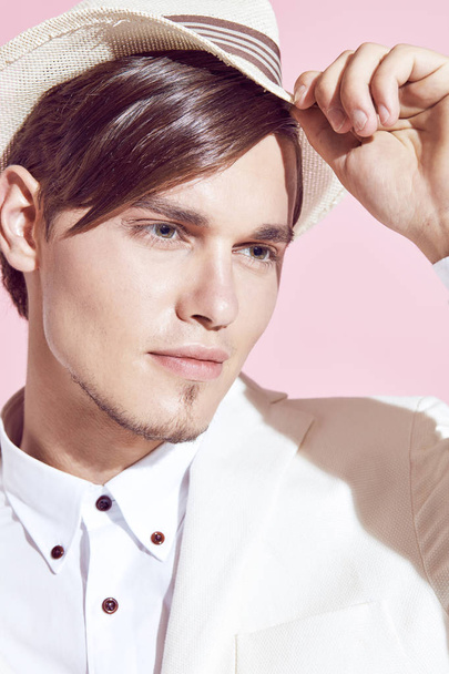 Closeup portrait of young handsome serious modern male model wearing white jacket, white shirt, grey pants and white hat posing in studio with light pink background - Foto, afbeelding