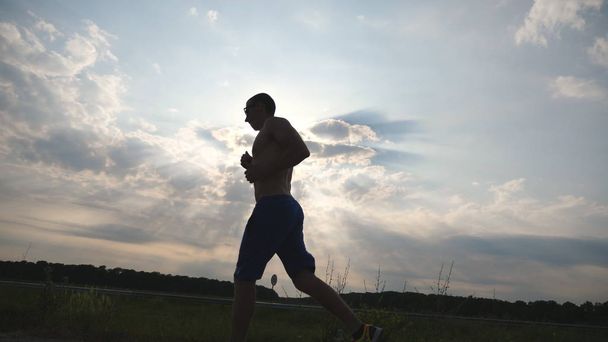 Σιλουέτα του μυϊκού Ανθρώπου τζόκινγκ στον δρόμο της χώρας κατά το ηλιοβασίλεμα. Ανδρική εκπαίδευση δρομέα για Μαραθώνα τρέξει έξω. Αθλητής που ασκεί και τρέχει εναντίον του γαλάζιου ουρανού. Αθλητισμός και ενεργός τρόπος ζωής αργή κίνηση - Φωτογραφία, εικόνα