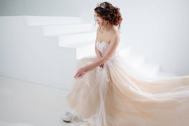 Bride sits on the stairs. Portrait of a beautiful girl in a wedding dress. Dancing Bride, white background. - Fotó, kép