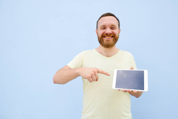 Счастливый улыбающийся красивый молодой человек с бородой показывает что-то на планшете изолированы на голубом фоне, яркий и легкий
 - Фото, изображение