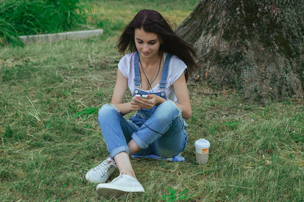 Młoda brunetka czarujący siedzi na trawie, słuchanie muzyki i patrząc na telefon - Zdjęcie, obraz