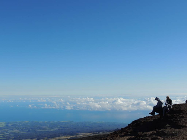 Nuori mies poika ja nainen tyttö teini istuu katselemassa lämpimiä ja kylmiä värejä auringonnousun yli Haleakalan kansallispuisto tulivuori Maui Havaiji USA pilvi kannen alapuolella näköalapaikka ja valtameren ja Pohjois-Maui
 - Valokuva, kuva