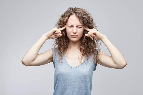 Primer plano de estudio de estresado infeliz joven mujer caucásica usando gris casual camiseta manteniendo los dedos en las sienes, sufriendo de dolor de cabeza severo, migraña o tratando de recordar algo
. - Foto, Imagen