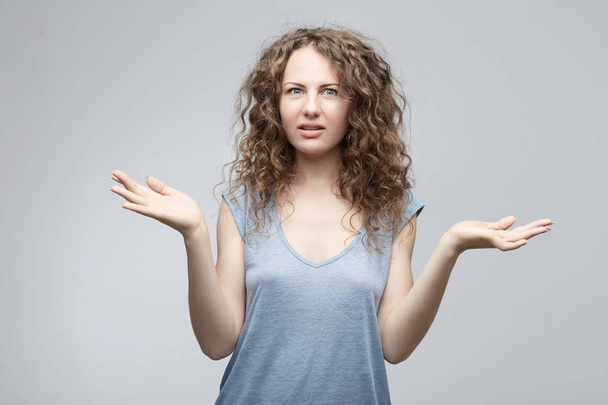 Unsure confused doubtful European woman with long curly hair in grey casual t-shirt shrugging shoulders in questioning gesture of uncertainty and keeping mouth opened,having confused clueless look. - Fotografie, Obrázek