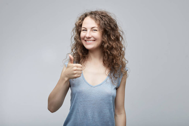 Ritratto di una giovane donna felice caucasica di successo che dà in camicia grigia casual mostrando pollice verso l'alto o come gesto, guardando e sorridendo fotocamera con espressione felice. Emozioni umane, espressioni facciali
. - Foto, immagini