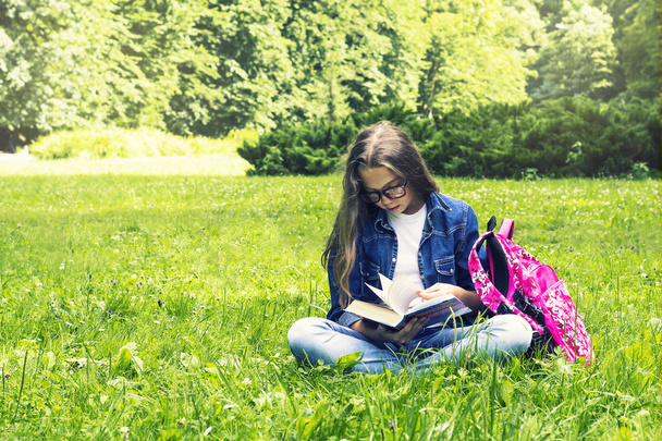 Beautiful blonde schoolgirl girl in jeans shirt reading a book on grass with a backpack in the park - Photo, image