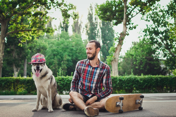 Young hipster man sitting on skateboard with funny siberian husky dog in cap  - Foto, Bild