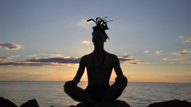 Silhouette eines Mannes mit indianischen Federmohawk-Accessoires auf dem Kopf, der bei Sonnenuntergang Yoga praktiziert - Filmmaterial, Video