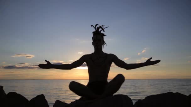Silhouette eines Mannes mit indianischen Federmohawk-Accessoires auf dem Kopf, der bei Sonnenuntergang Yoga praktiziert - Filmmaterial, Video