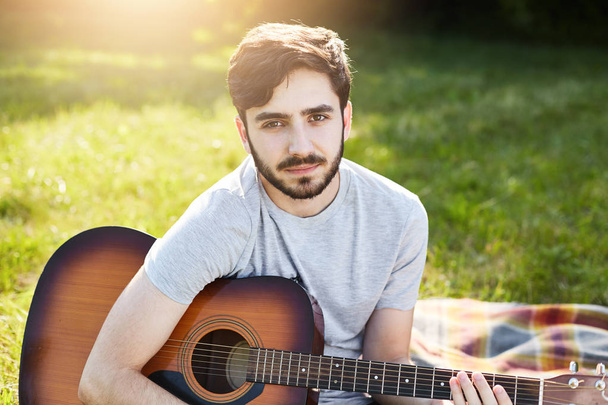 Young bearded male guitarist in casual T-shirt sitting with musical instrument on green grass, playing different songs, looking at camera with serious look, resting at nature. People, hobby, music - Foto, Bild