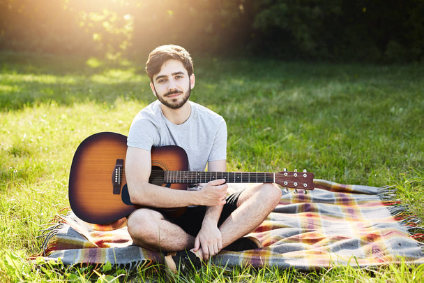Restful bearded guy sitting crossed legs at beautiful meadow, holding guitar, trying to learn how to play it, feeling inspiration and relaxation after hard work or studying. Young musician outdoor - Фото, зображення