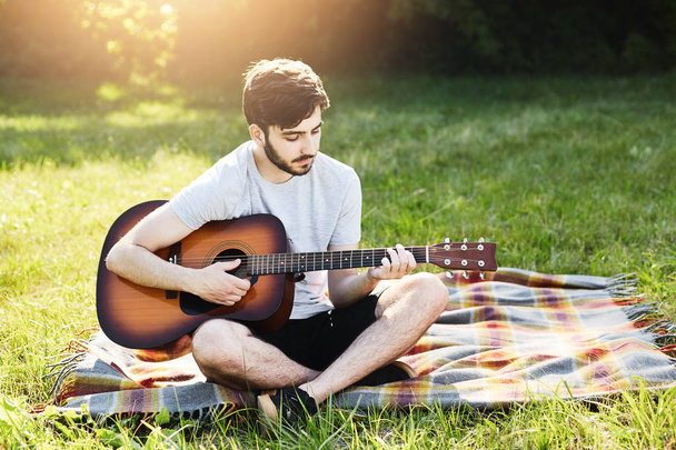 Portrait of attractive stylish bearded guy with guitar sitting crossed legs on green grass, playing musical instrument, enjoying calm atmosphere. Talented boy demonstrating his talent while resting - Photo, image