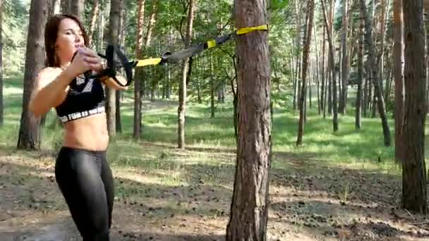 Beautiful, athletic, sexy young woman, coach, instructor, performs exercises, doing exercises with fitness trx system, TRX suspension straps. In pine forest, in summer, in sun rays. - Materiał filmowy, wideo