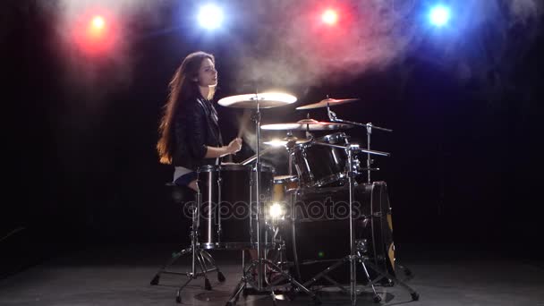 Girl plays the drums and smiles. Black smoke background. Red blue light from behind. Side view - Záběry, video
