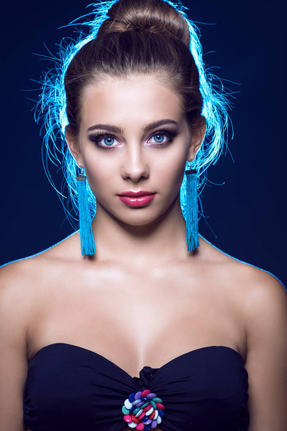 Nahaufnahme Porträt eines extrem schönen jungen Mädchens mit ausdrucksstarken blauen Augen und perfektem Make-up, das schwarzes trägerloses Top und blaue Quasten-Ohrringe trägt, Babyhaare um ihren Kopf, die im Gegenlicht leuchten - Foto, Bild