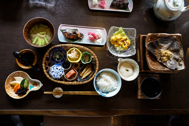 Japanese ryokan breakfast dishes including cooked white rice, grilled fish, fried egg, soup, mentaiko, pickle, seaweed, hot plate, other side dishes and green tea on wooden table - 写真・画像