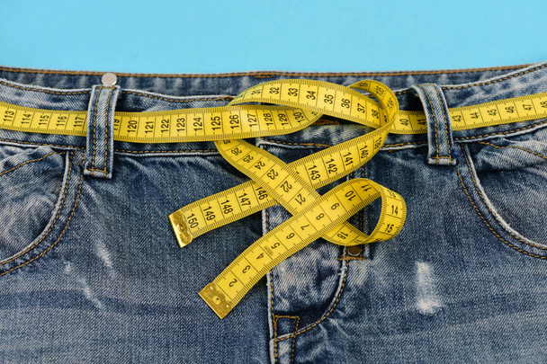 Jeans with yellow measure tape as belt on blue background - Foto, afbeelding
