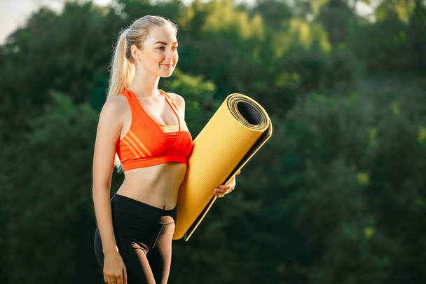 A young blonde in a red top and black pants holds a mat for a fountain in the nature. A woman prepares for gymnastics - Foto, Imagen