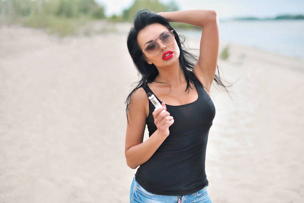 Young sexy woman with big breast vaping e-cigarette near blue water on the beach - Photo, image