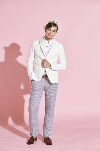 Young beautiful serious modern man wearing white jacket, white shirt, grey pants and white hat standing and posing in studio with light pink background - Photo, image