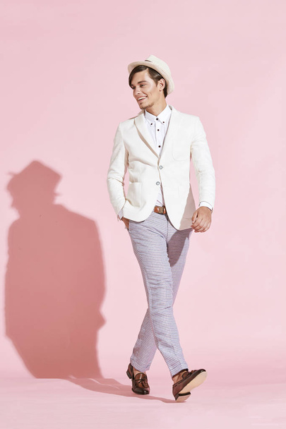 Young beautiful serious modern man wearing white jacket, white shirt, grey pants and white hat posing in studio with light pink background - Foto, Imagen