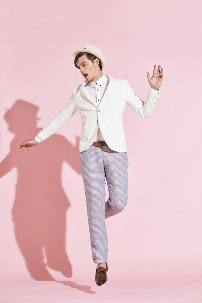 Jovem bonito sério homem moderno vestindo jaqueta branca, camisa branca, calças cinza e chapéu branco pulando e posando com a boca aberta em estúdio com fundo rosa claro
 - Foto, Imagem